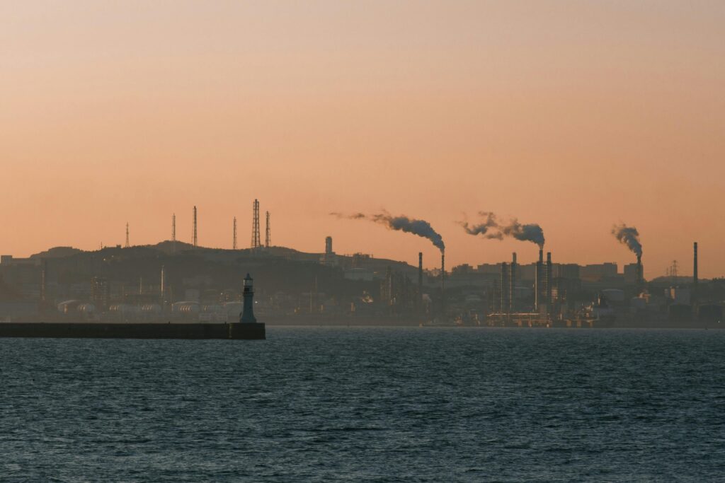 Factory skyline emitting smoke over the ocean at sunset, under an orange sky.